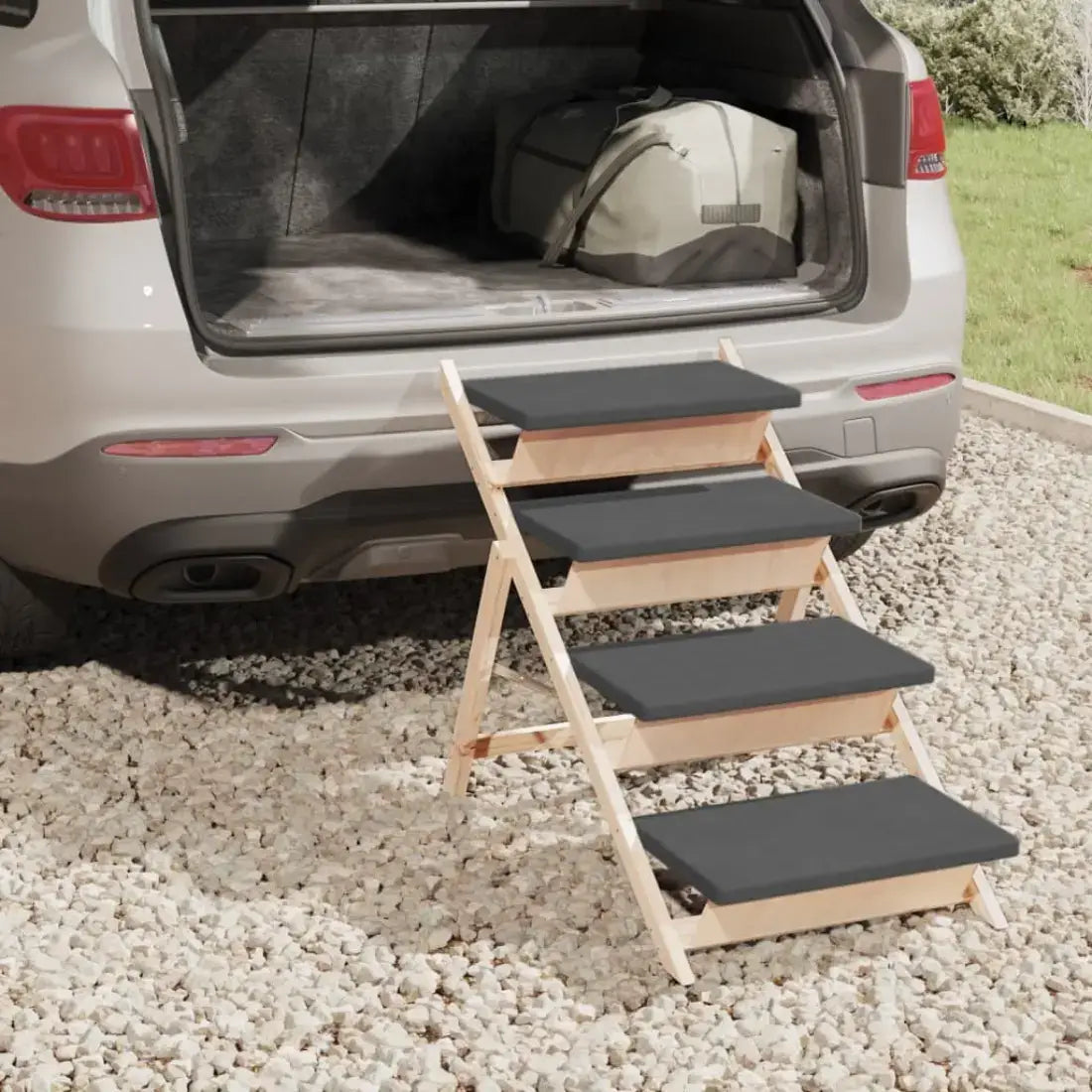 A wooden pet step ladder with black rubberized steps is positioned in front of an open car trunk on a gravel surface outdoors.