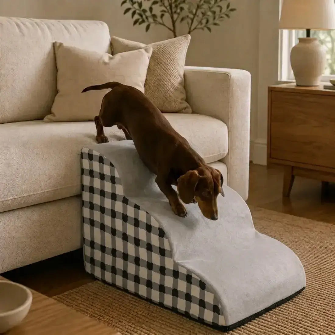 A brown dachshund is climbing a two-step pet ramp with a black and white gingham checkered side panel and a light gray top surface, positioned next to a beige sofa in a living room setting.
