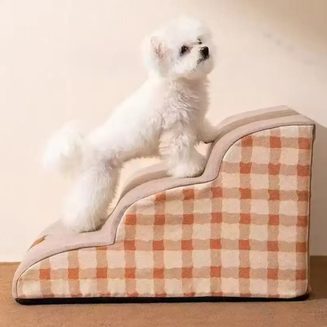 A small white dog stands on a padded indoor pet staircase with a beige and orange gingham patterned fabric, photographed against a plain light wall.