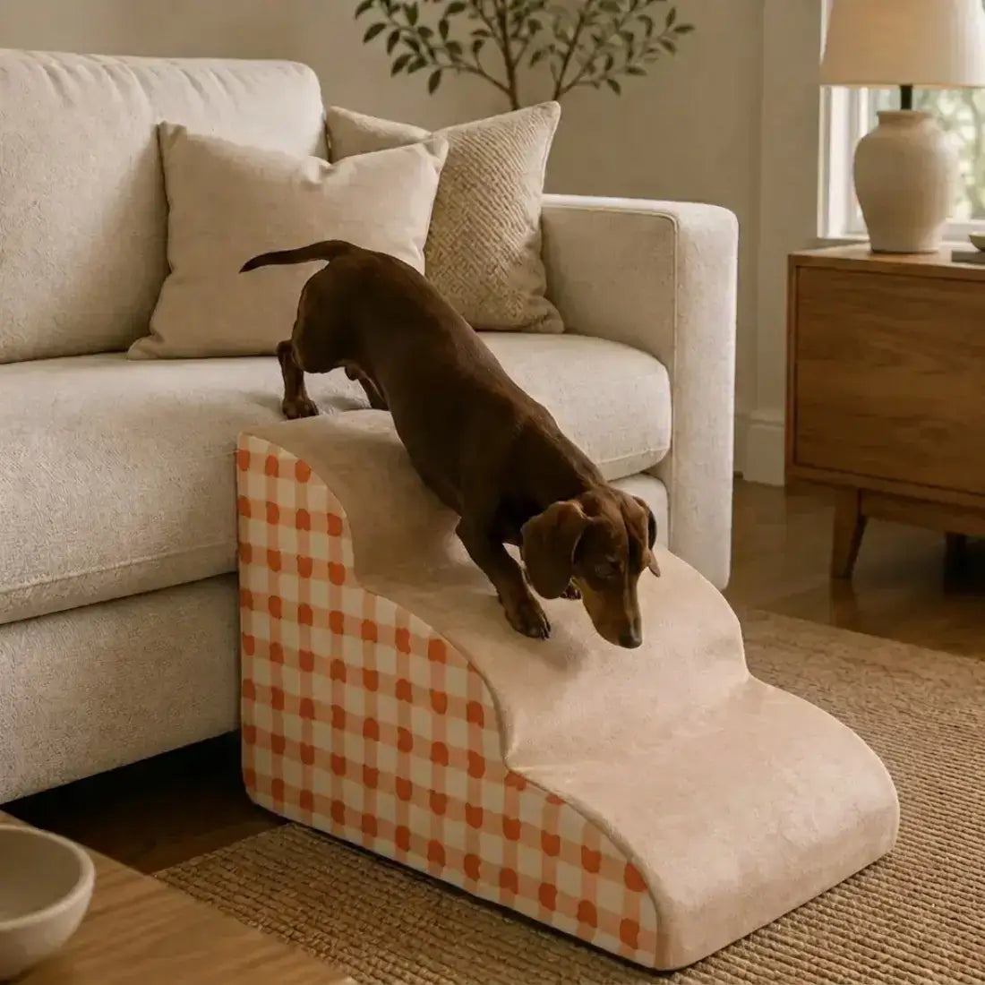 A dachshund dog is climbing a small step pet ramp positioned next to a light beige sofa in a cozy living room setting. The ramp features a cream-colored plush top and a side panel with an orange and cream gingham checkered pattern.