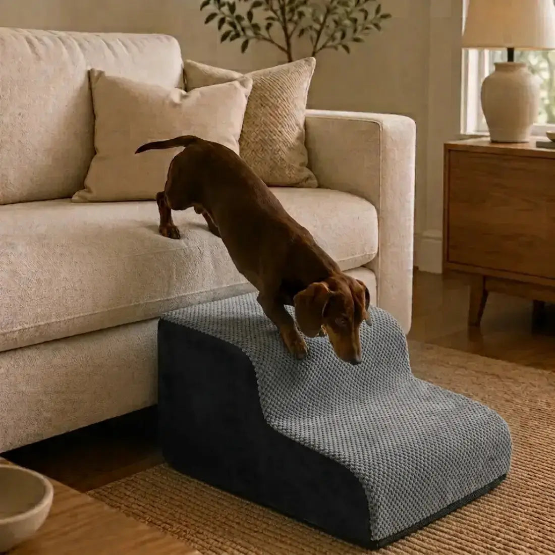 A brown dachshund dog is climbing a two-step pet ramp with a textured blue-gray top surface and a dark charcoal base, positioned in front of a beige sofa in a living room setting.